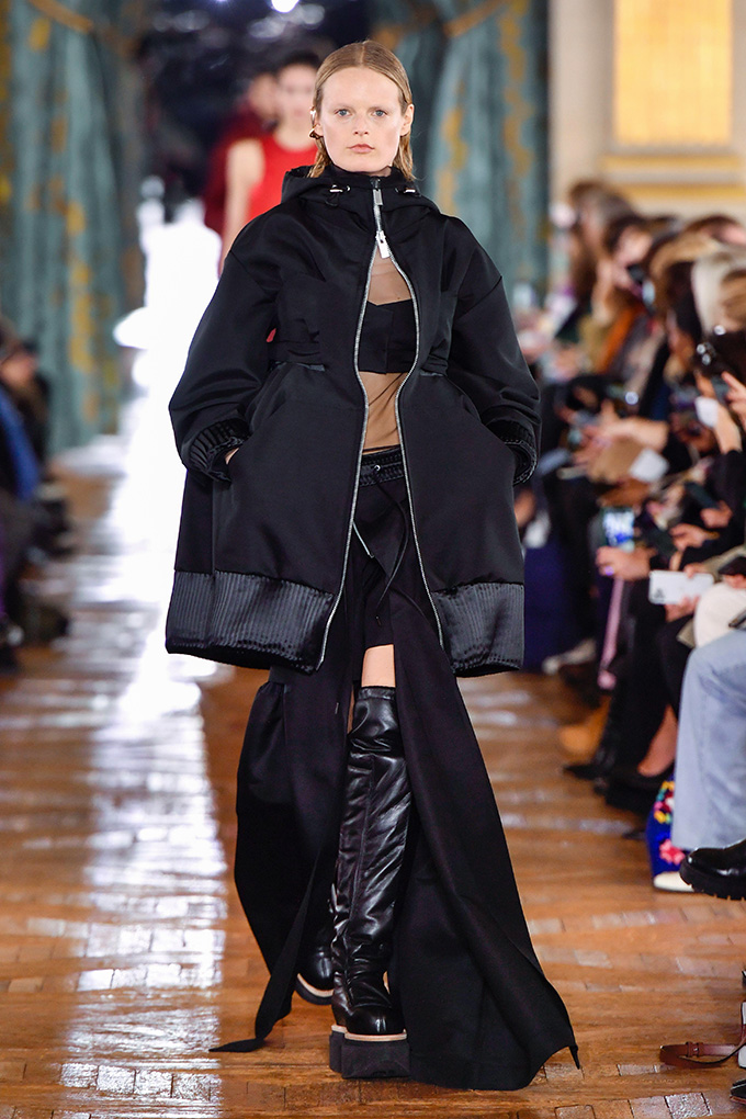 Model Hanne Gaby Odiele walks a track with blonde hair sliding rear and dressed in a large black jacket, a top top with a large black ribbon beyond the chest, a black skirt with a high cut boots of thighs and dense black thigh leather boots. Odiel is interseks.